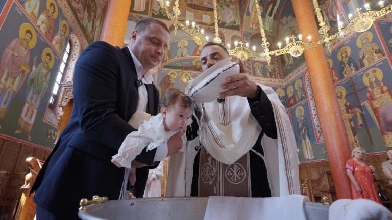 Sveštenik preliva vodom bebu na krštenju u crkvi sa freskama